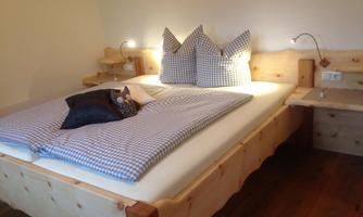 A cozy bedroom with a large wooden bed and checkered pillows. On the nightstand, there are reading lamps and personal belongings.