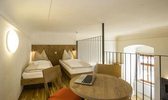 A modern room with two single beds and a small table. The room has large windows and plenty of natural light.