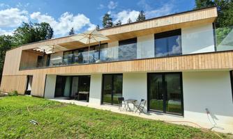 A modern house with a wooden facade and large windows. In front of the house, there is a small garden with a table and chairs.