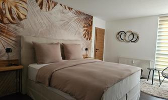 A modern bedroom with a large bed and soft colors. The wall is decorated with a plant pattern, and there is a bright atmosphere from the natural light.