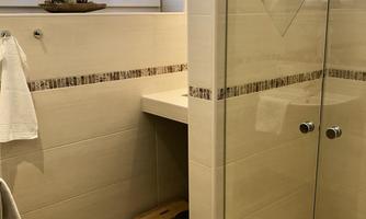 A modern bathroom with a glass shower wall and a small wooden stool. The walls are brightly tiled and there are windows that let in natural light.