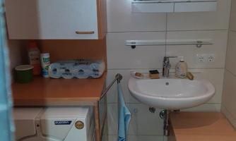 A small bathroom with a washing machine and a sink. A mirror is hanging on the wall and there is a wooden cabinet as well as a waste bin.