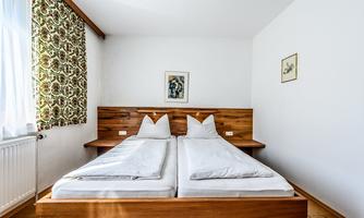 A simple bedroom with two wooden single beds. Curtains hang next to the windows and some pictures are decoratively placed on the wall.