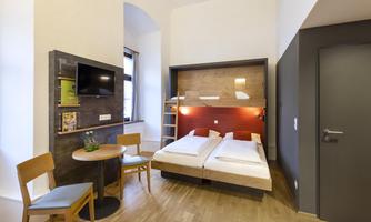 A modern hotel room with two single beds and wooden flooring. There is a small table, chairs, and a television on the wall.