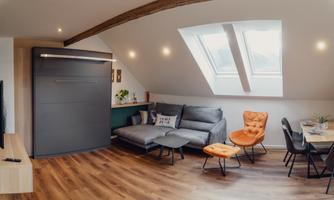 A modern penthouse with a cozy living room and many windows. The furniture consists of a sofa bed, a dining table, and a striking chair.