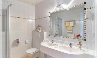 A modern bathroom with a large shower and a double sink. On the sink, there is a small vase with a red flower.