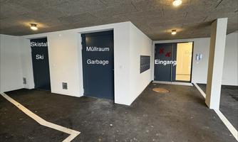 A parking lot with two doors leading to the ski storage and trash room. The entrance to a building is also visible.
