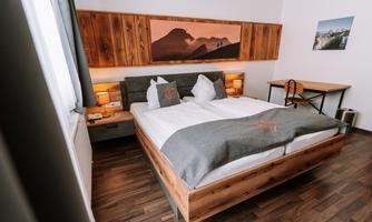 A modern bedroom with a large double bed and wooden details. A picture of mountains hangs on the wall, and a desk stands next to the window.