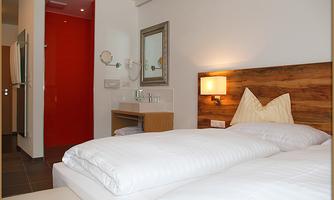 A modern bedroom with two single beds and a wood-paneled headboard. In the background, you can see a bathroom with a striking red wall.