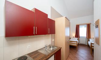 A modern kitchenette with red cabinets and a sink. In the background, two single beds and a window with curtains are visible.