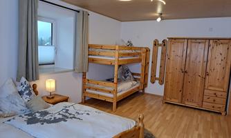 A cozy bedroom with two wooden beds and a bunk bed. The room has a bright atmosphere and a large wardrobe.
