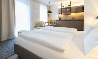 A modern bedroom with a double bed and light curtains. In the background, there is a small kitchenette and dining area.