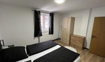 A simple bedroom with two single beds and black bedspreads. The walls are white, and there is a closet as well as a wooden dresser in the room.