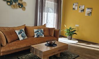 A cozy living room with an orange sofa and a wooden table. Decorative elements hang on the wall, and there is a plant in the corner.