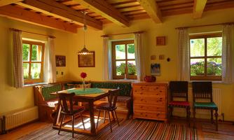 A cozy room with a wooden table and chairs. The walls are painted yellow, and there are windows overlooking greenery.