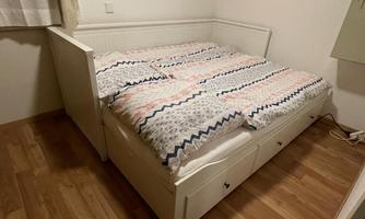 A bed with a white frame and colorful bedspreads. The floor is made of light wood and there is a window with curtains.
