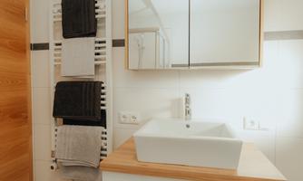 A modern bathroom with a white sink and a wooden shelf. Towels are hanging on the wall and a mirror is above the sink.