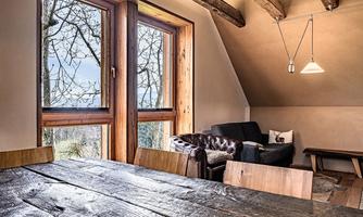 A cozy living space with wooden details, a large table, and a comfortable sofa. Large windows offer a view of the nature outside.