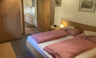 A simple bedroom with a double bed and red checkered blankets. In the background, an open door to the kitchen is visible.