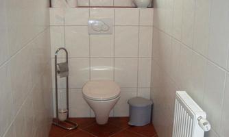 A small bathroom with a toilet, a window, and a radiator. The walls are light and tiled, the floor is brown.