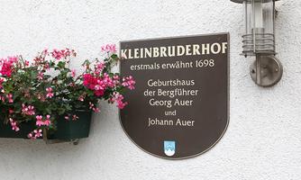 A sign with the inscription "Kleinbruderhof" and information about the birthplace of the mountain guides Georg Auer and Johann Auer. Next to it, flower pots with pink blossoms are hanging.