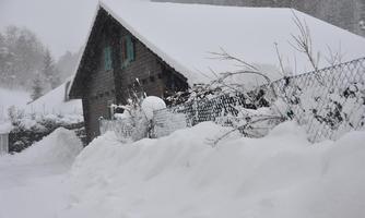 A snow-covered house in a wintry landscape. High snow drifts and thin branches are visible.