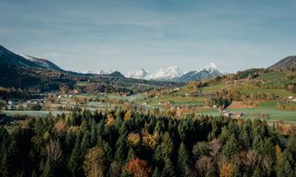 A picturesque landscape with green meadows and colorful foliage in the foreground. In the background, snow-covered mountains rise under a clear sky.