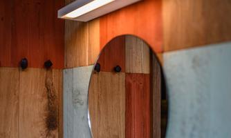 A round mirror hangs in front of a rustic wall made of wooden panels in warm colors. The lighting above the mirror creates a pleasant atmosphere.