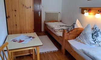 A simple room with two single beds, a table, and a wooden wardrobe. There is a quiet and cozy atmosphere.