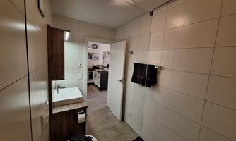 A modern bathroom with tiled walls and a sink. In the background, a kitchen is visible.