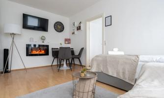 A modern living room with a cozy sofa and a dining table. A television hangs on the wall above an electric fireplace.