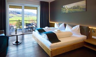 A modern room with a large bed and a view of the countryside. There is a small table and a balcony with chairs.