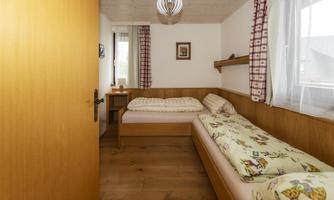 A cozy bedroom with two single beds and wooden shelves. Bright walls and windows create a friendly atmosphere.