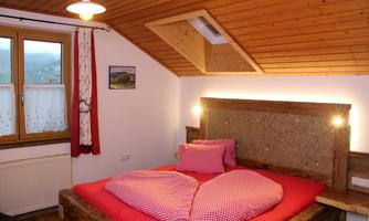 A cozy bedroom with a wooden bed and red heart-shaped pillows. There is a window with a view of the countryside and a rustic wooden ceiling.