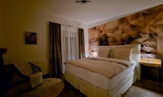 A cozy bedroom with a large bed and soft lighting. The walls are decorated with a warm, natural pattern.