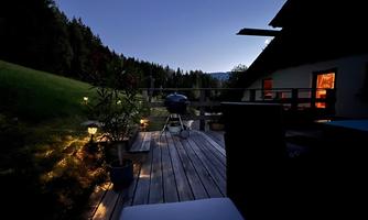 A cozy balcony at night, surrounded by trees and soft lighting. In the background, there is a grill and the view of nature is visible.