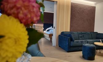 A modern room with a blue sofa and a small table. In the background, a bed is visible, surrounded by warm wood paneling.