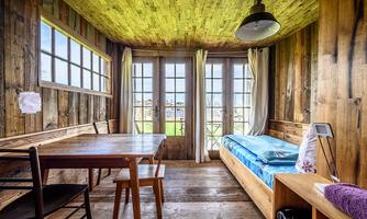 A cozy room with wooden paneling and large windows. A table, a chair, and a bed are present in the room.