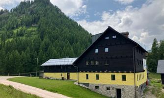 A modern building with a dark brown and yellow facade stands at the foot of a green hill. The sky is partly cloudy and the landscape is surrounded by trees.