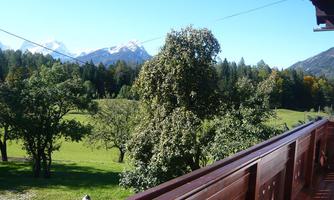 A picturesque view from a balcony overlooking green meadows and wooded mountains. In the background, snow-covered peaks can be seen.