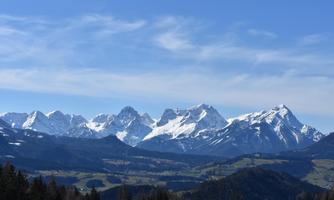 A majestic mountain landscape with snow-covered peaks and a clear blue sky. In the foreground, green valleys and forests extend.