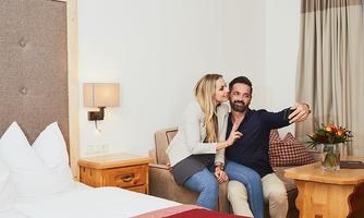 A happy couple takes a selfie in a cozy room. In the background, there is a bed and a flower arrangement.