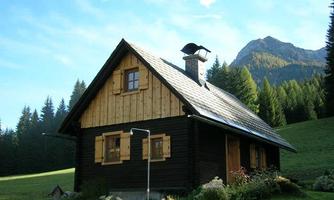 A cozy wooden house in nature, surrounded by green meadows and tall trees. In the background, gentle mountains can be seen.