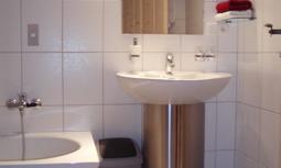 A modern bathroom with a round sink and a bathtub. The walls are tiled, and there are towels as well as a waste bin.