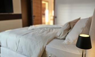 A cozy bedroom with a large bed and a modern bedside table. Next to the bed is a decorative lamp on a wooden tray.