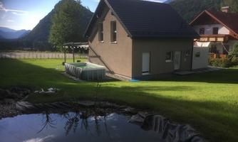 A modern house in a natural environment with a view of the mountains. In the foreground, there is a pond and a pool in the garden.
