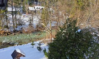 A snowy landscape painting with a clear river, surrounded by trees. In the background, a few huts can be seen.