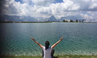 A person is sitting by the shore of a clear lake and spreading their arms. In the background, mountains and a blue sky with clouds can be seen.