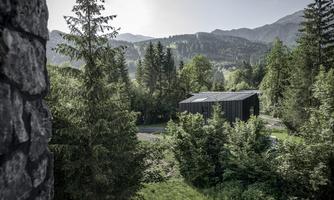 A modern house stands surrounded by trees and mountains. The landscape is green and peaceful.