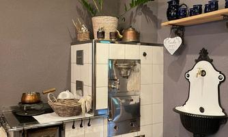 A cozy kitchen with a traditional stove and wooden shelves. The walls are painted in a warm gray shade, and there are decorations like plants and dishes.
